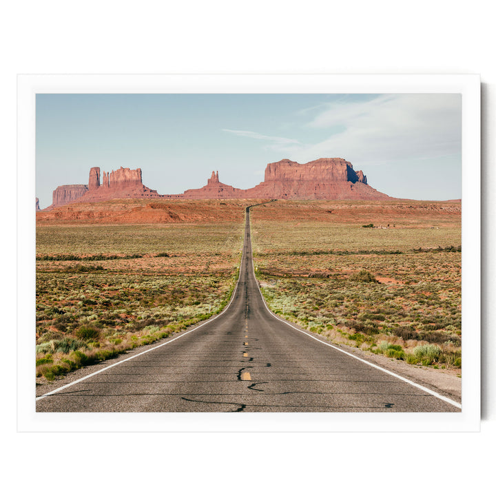 A long, straight road stretches out into the distance, leading towards a series of red rock formations in the background.
