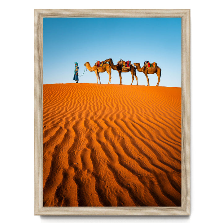 A person in a blue robe leads three camels across a vast, undulating sand dune under a clear blue sky.
