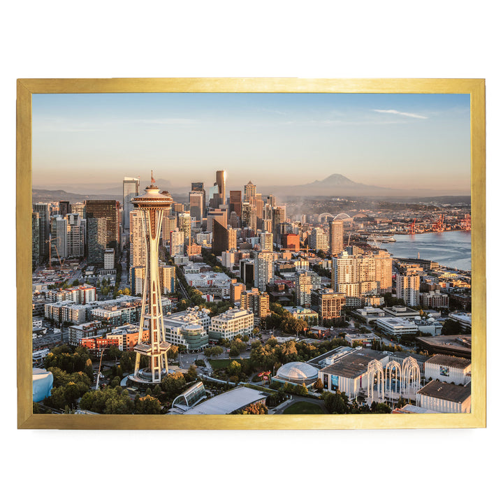 a cityscape with a tall tower, likely a landmark, surrounded by a variety of buildings and skyscrapers. The city appears to be located near a body of water, with a mountain visible in the distance. The sky is clear and blue, suggesting a bright and sunny day. The image is framed within a gold border, giving it a polished and professional appearance.
