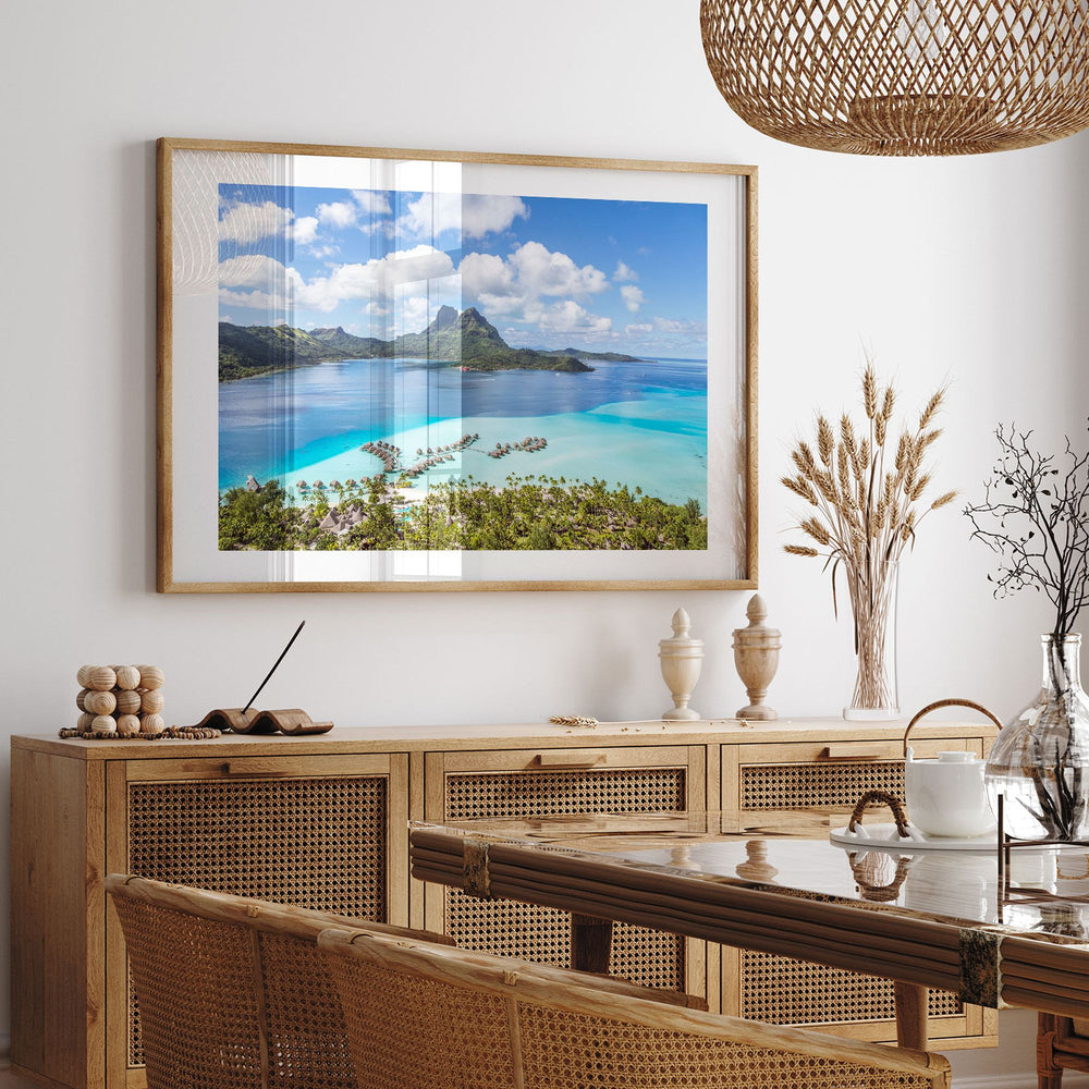a large framed picture of a tropical beach scene hanging on a wall above a wooden cabinet with drawers. The cabinet has a glass top and is situated in a dining area with a table and chairs. The overall setting appears to be a cozy and inviting space, with a focus on the natural beauty of the beach landscape.