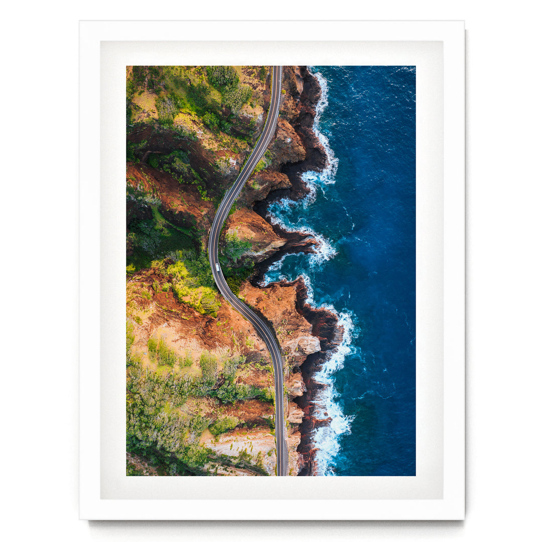 An aerial view of a winding road cutting through a rugged coastline with cliffs, water, and vegetation.