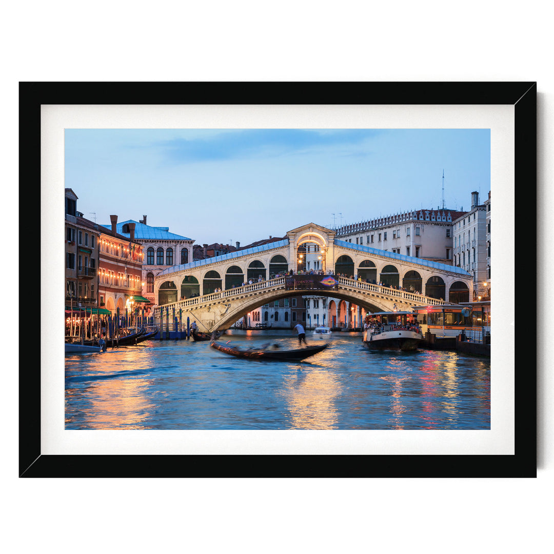 a picturesque scene of the Rialto Bridge in Venice, Italy, with its distinctive white stone arch spanning the Grand Canal. The bridge is illuminated at dusk, creating a warm and inviting atmosphere. The canal is filled with boats, adding to the lively and bustling ambiance of the scene. The image is framed within a black border, highlighting the architectural beauty of the bridge and the surrounding buildings.