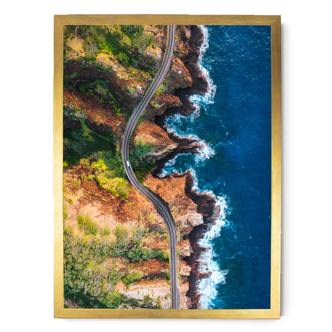 The image is an aerial view of a winding road cutting through a rugged, mountainous landscape with cliffs and the ocean.