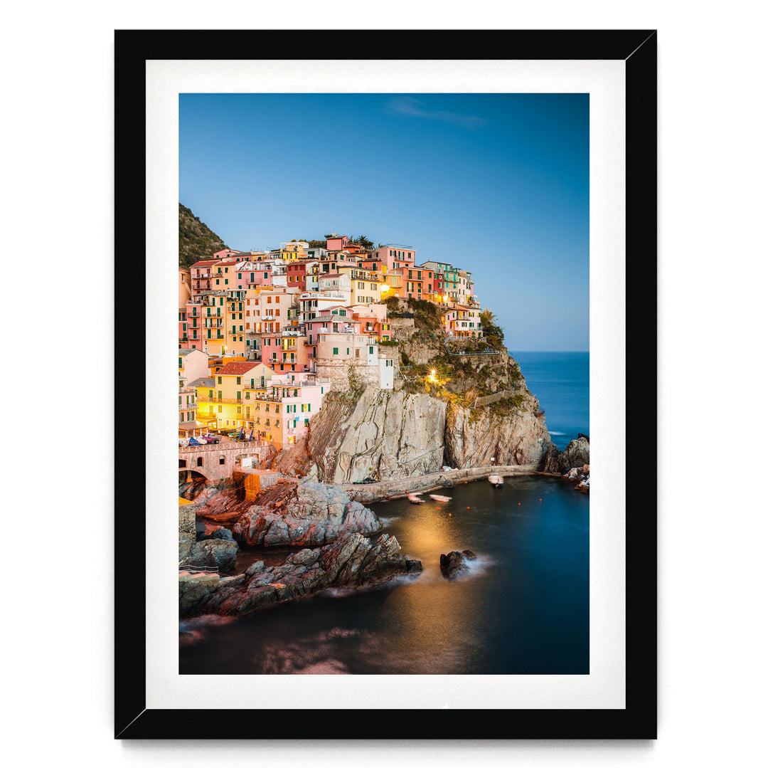 A picturesque coastal town nestled on a cliff, with its buildings illuminated by warm lighting against a clear blue sky.