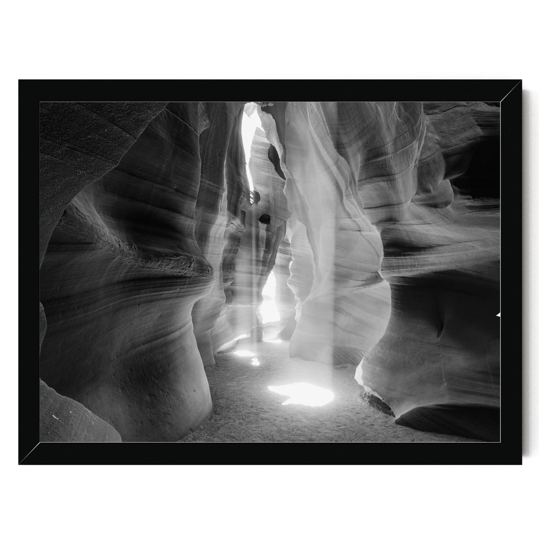 A black and white image of a narrow passage in a rock formation, with sunlight streaming through the opening and casting shadows on the walls.