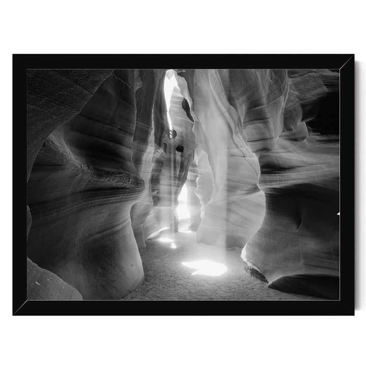 A black and white image of a narrow passage in a rock formation, with sunlight streaming through the opening and casting shadows on the walls.
