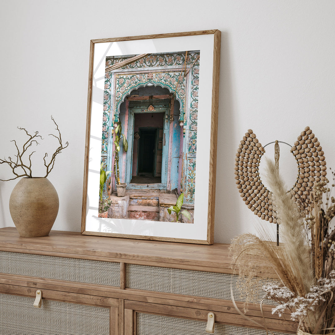 a wooden shelf with a framed picture of an ornate doorway, a vase with dried branches, and a woven basket with dried flowers.