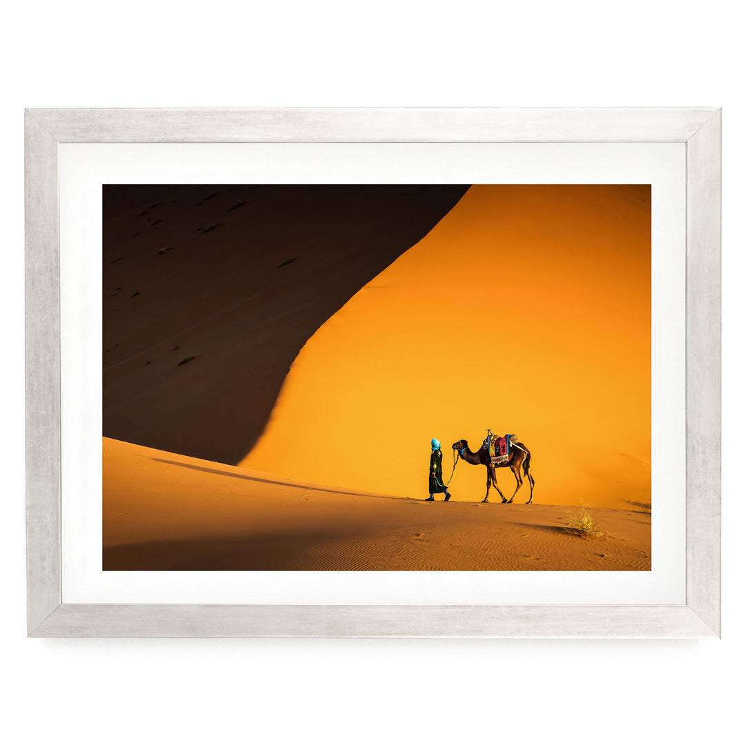 A person in a green outfit is walking alongside a camel in a desert setting, with a large sand dune in the background.