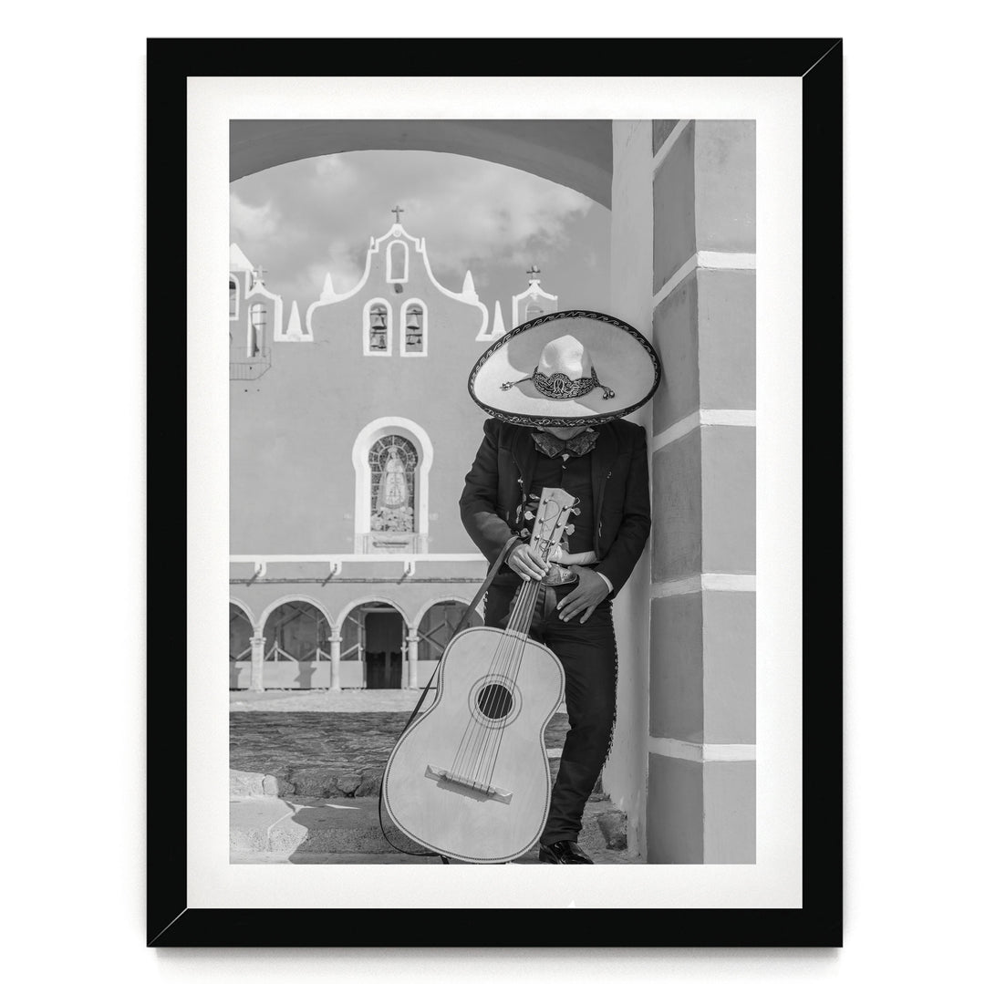 A person wearing a sombrero and holding a guitar stands in front of a building with arched windows and a cross on top.