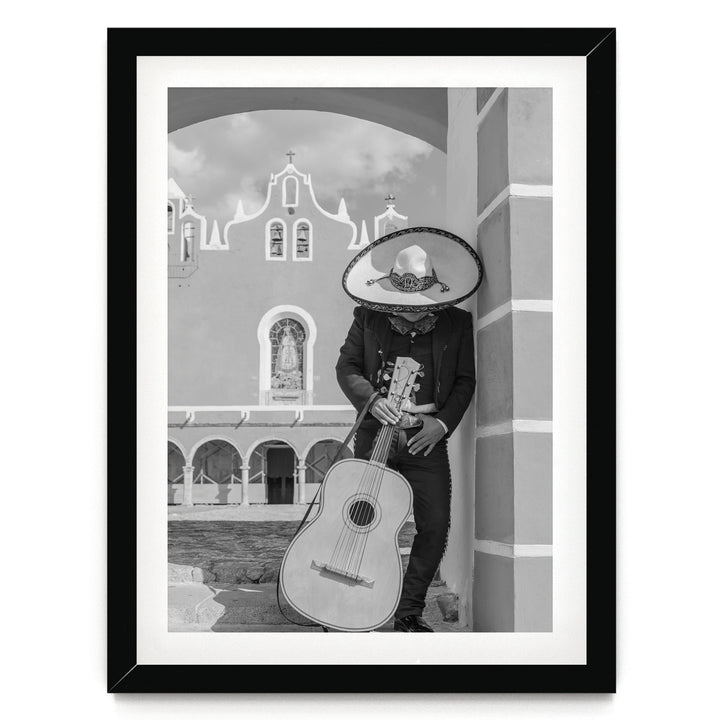 A person wearing a sombrero and holding a guitar stands in front of a building with arched windows and a cross on top.