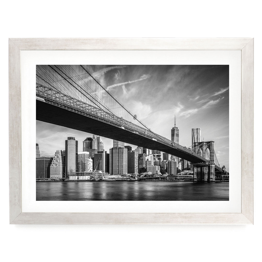 A black and white photograph of the Brooklyn Bridge spanning the East River, with the city skyline of New York City visible in the background.