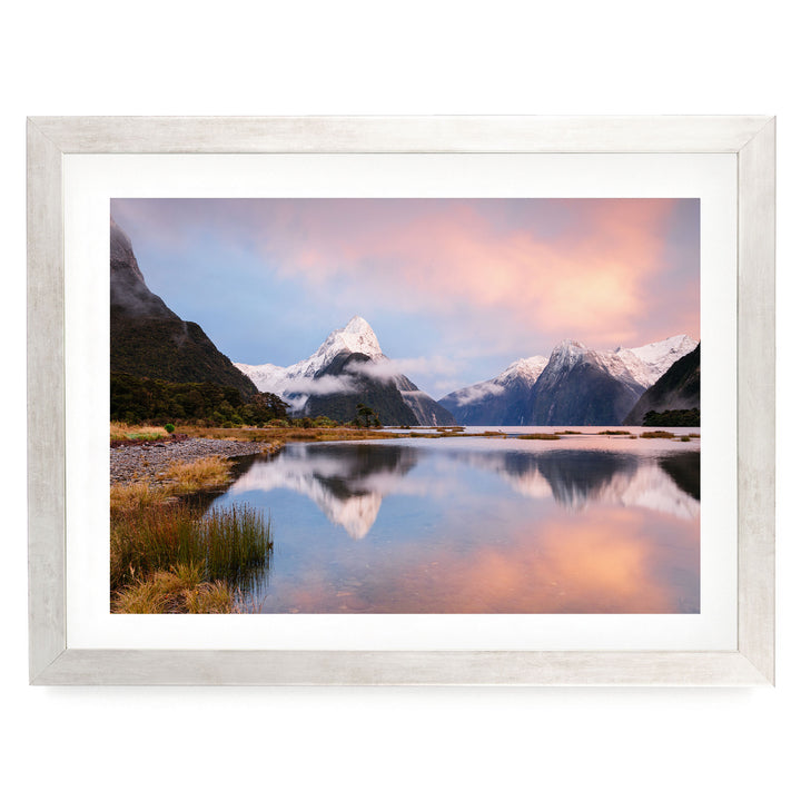 a serene landscape with a calm lake reflecting the surrounding mountains and sky. The mountains are covered in snow, and the sky is a beautiful gradient of pink and orange, creating a stunning backdrop for the scene.