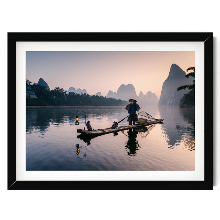 A person stands on a long wooden boat, paddling across a calm body of water surrounded by mountains and trees.