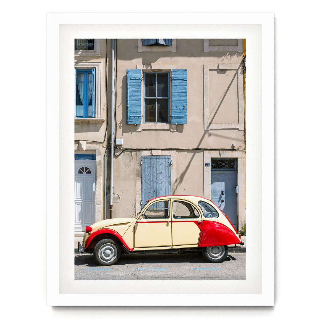A vintage yellow and red car is parked on a street in front of a building with blue shutters and a white door.