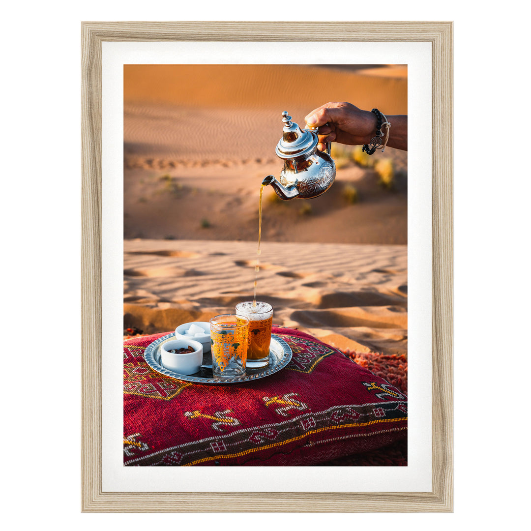 A hand pours tea from a silver teapot into a glass, with a tray of tea and snacks on a red and gold patterned cushion, set against a desert landscape.