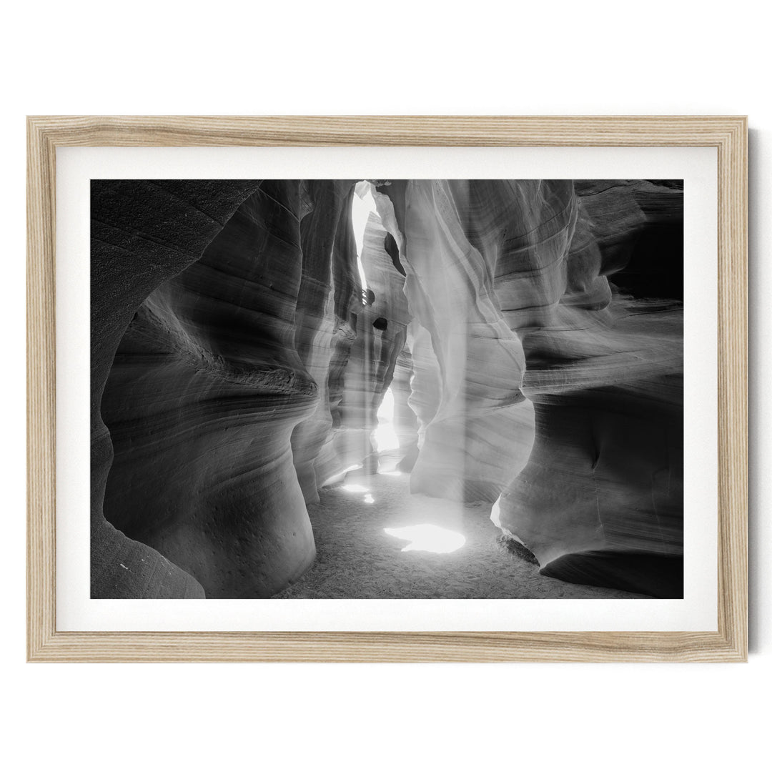 A black and white photograph of a narrow passage in a canyon, with sunlight streaming through the walls and creating a dramatic, ethereal atmosphere.