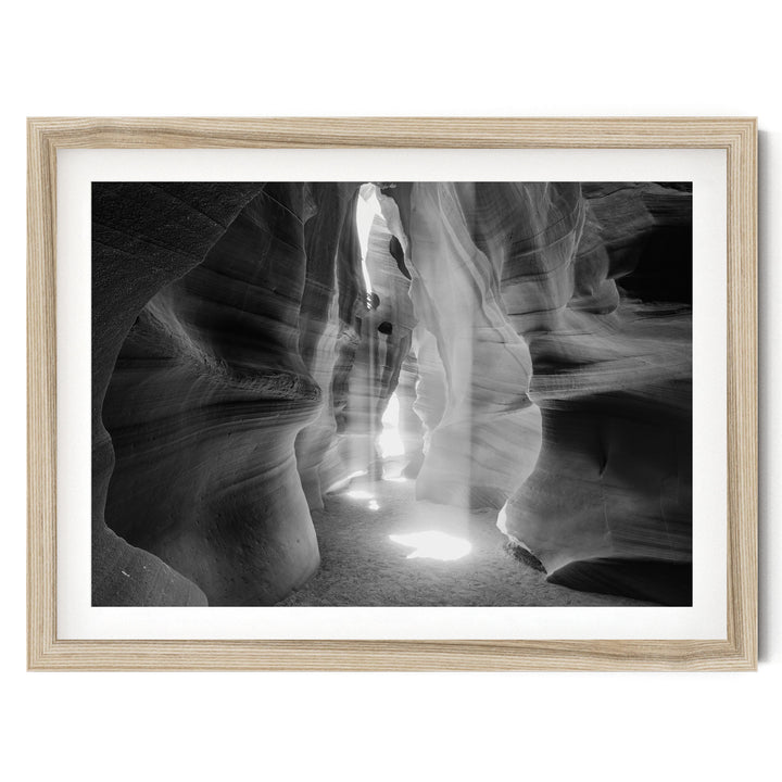 A black and white photograph of a narrow passage in a canyon, with sunlight streaming through the walls and creating a dramatic, ethereal atmosphere.