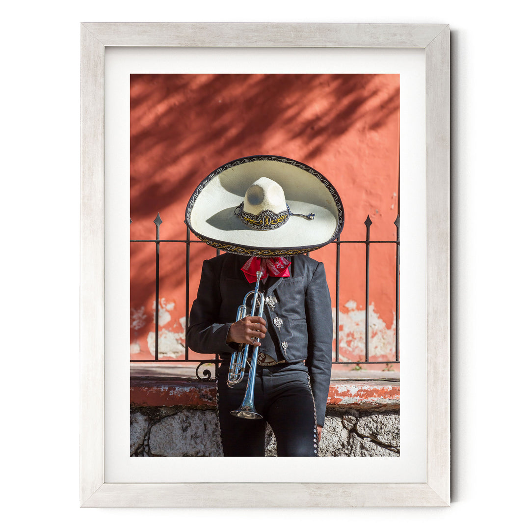 A person wearing a sombrero and a black jacket is playing a trumpet in front of a red wall.