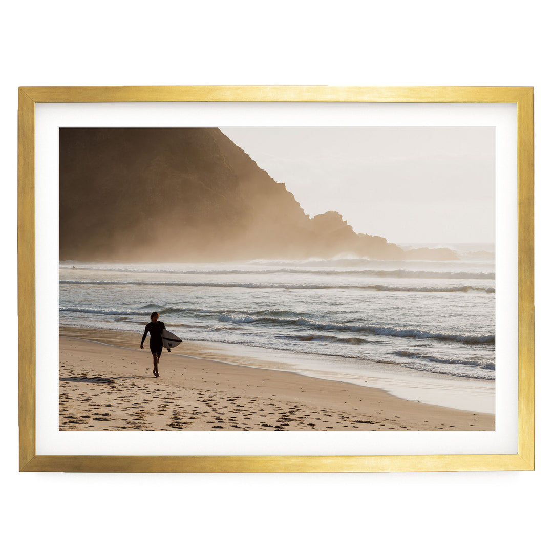 A surfer walks along the beach, carrying a surfboard, with a rocky cliff in the background.