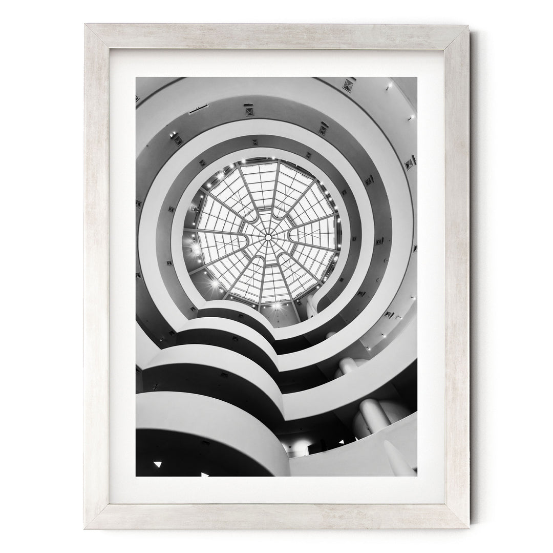 A black and white photograph of a spiral staircase with a large circular skylight at the top, creating a striking visual effect.