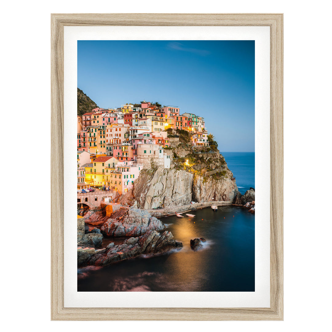A picturesque coastal town nestled on a cliff, with its buildings illuminated by warm lighting against a clear blue sky.
