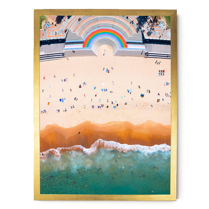 The image is an aerial view of a beach with a crowd of people enjoying the sunny day. The beach has a mix of light and dark colors, with the water appearing a vibrant shade of green. The sky above is a clear blue, and there is a rainbow arching across the scene. The image is framed within a gold border, giving it a decorative and artistic appearance.