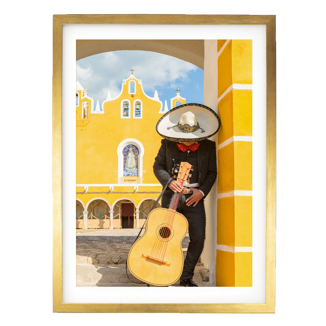 A person in a sombrero and mariachi outfit is playing a guitar in front of a yellow building with arched windows and a cross on top.