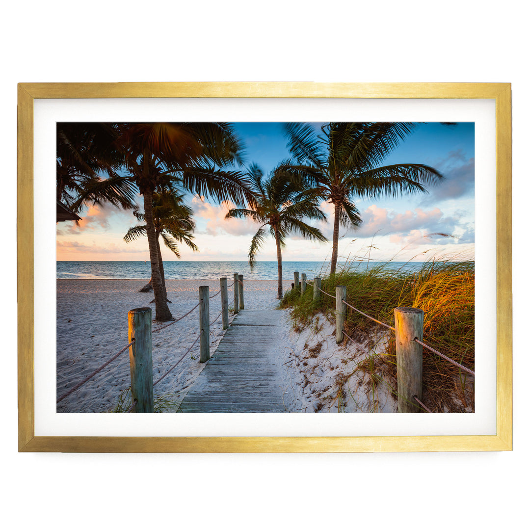 A wooden walkway leads to a sandy beach, with palm trees and a colorful sunset sky in the background.