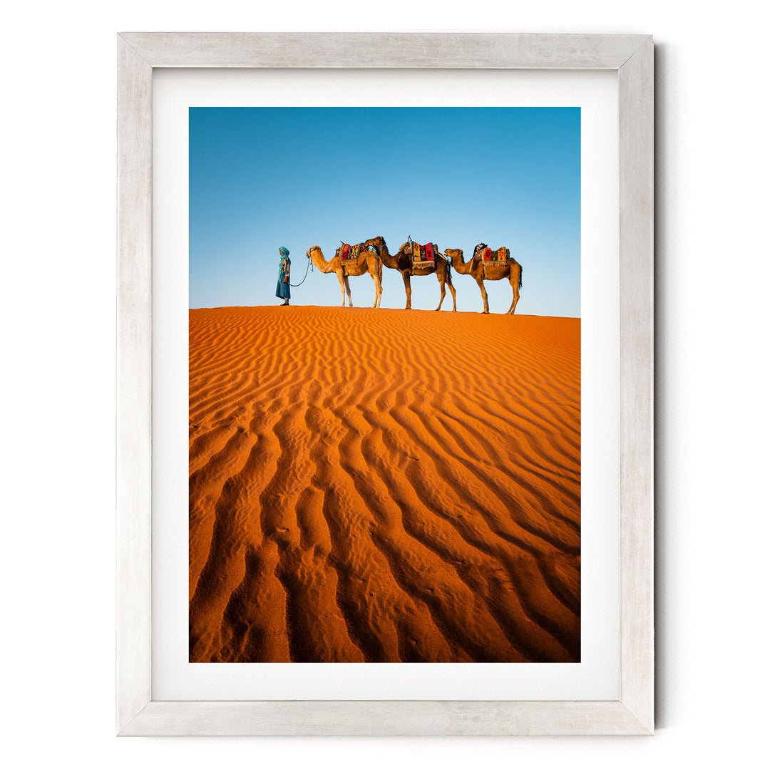 A person in a blue robe leads three camels across a vast, undulating sand dune in a desert landscape.