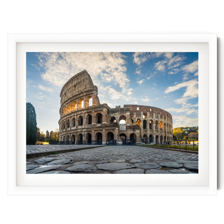 the Colosseum, a famous ancient amphitheater in Rome, Italy. The Colosseum has a distinctive oval shape and is surrounded by a stone courtyard. The sky above is blue with some clouds, creating a picturesque scene. The image is framed and appears to be a photograph of the Colosseum.