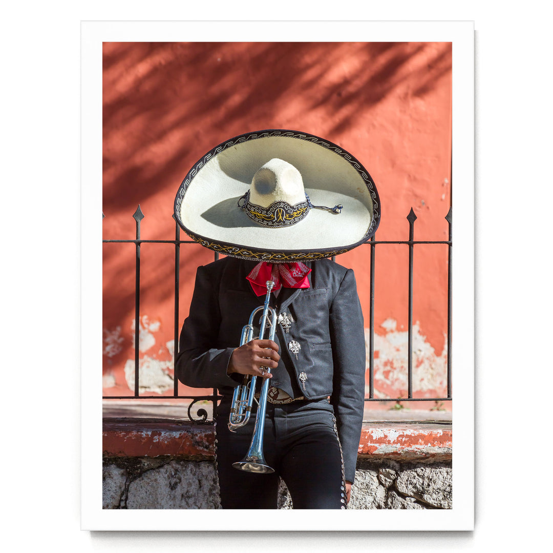 A person wearing a sombrero and traditional Mexican attire is playing a trumpet in front of a red wall.