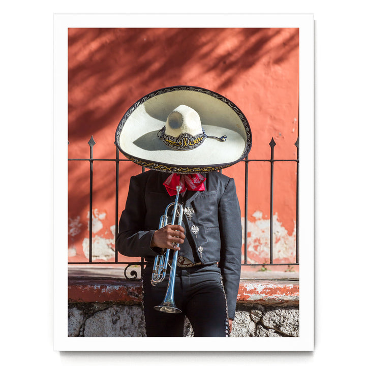 A person wearing a sombrero and traditional Mexican attire is playing a trumpet in front of a red wall.