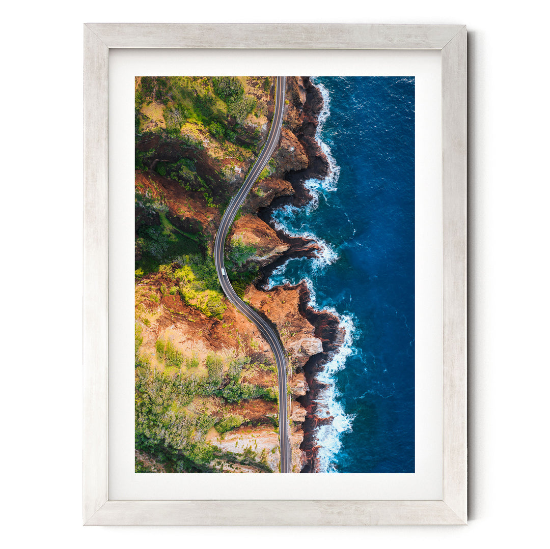 The image is an aerial view of a winding road cutting through a rugged coastline with cliffs and crashing waves.