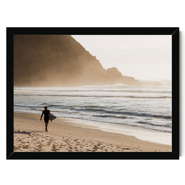 A person in a wetsuit walks along the beach, carrying a surfboard, with a rocky cliff in the background.