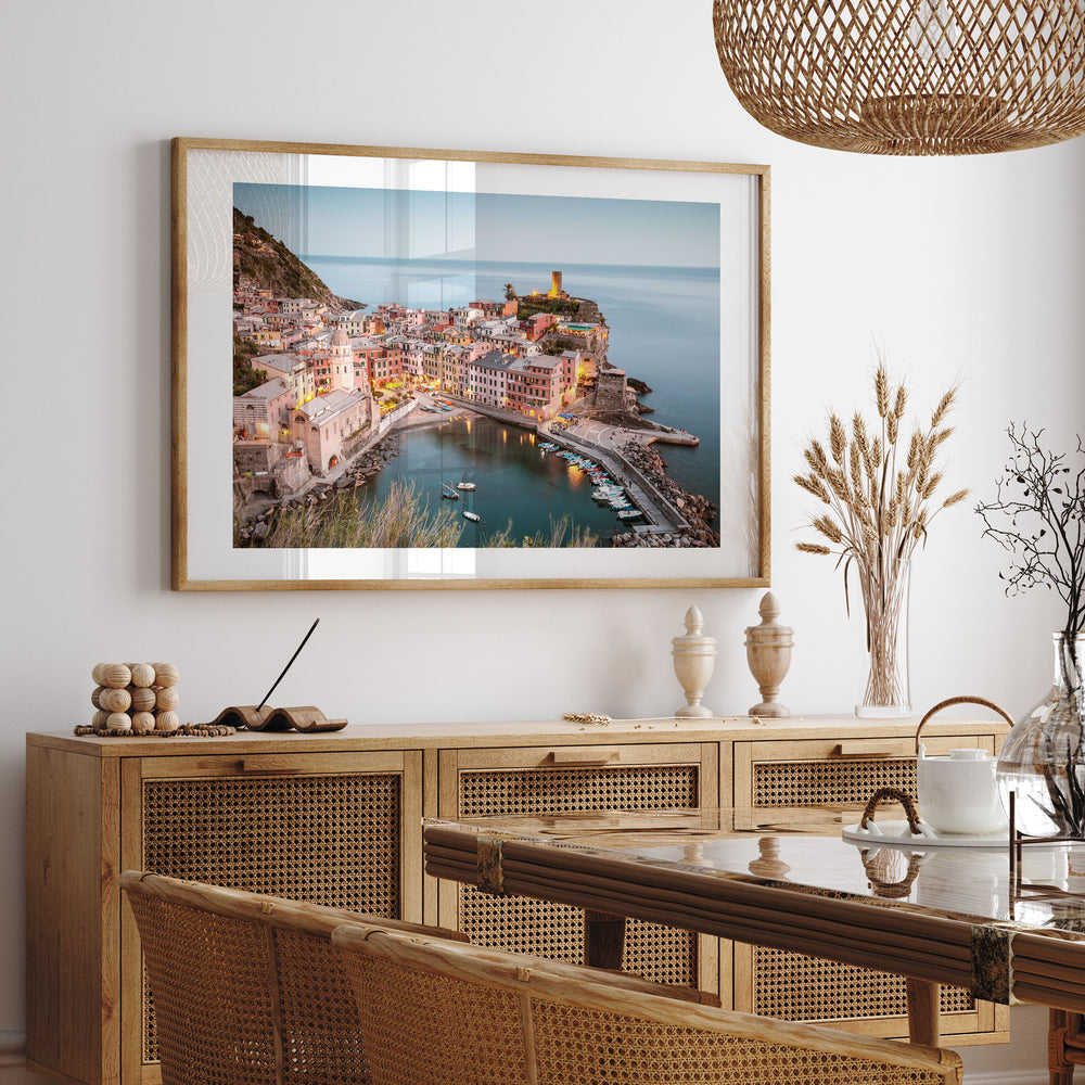 a large framed picture of a coastal town with a harbor, hanging on a wall above a wooden cabinet and a dining table.