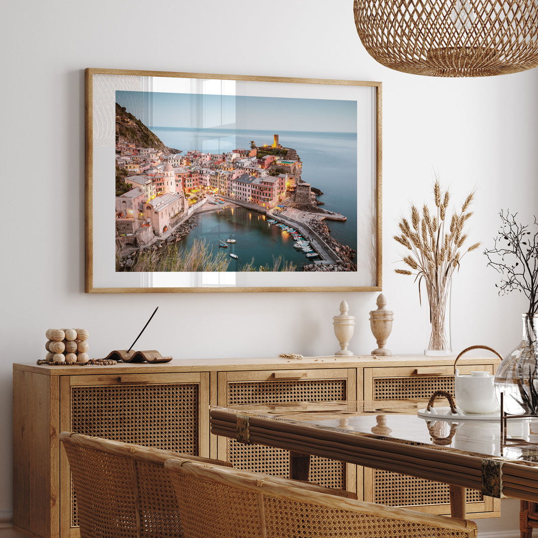 a large framed picture of a coastal town with a harbor, hanging on a wall above a wooden cabinet and a dining table.