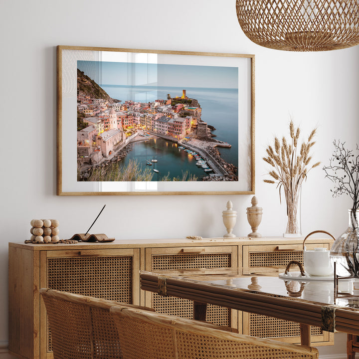 a large framed picture of a coastal town with a harbor, hanging on a wall above a wooden cabinet and a dining table.