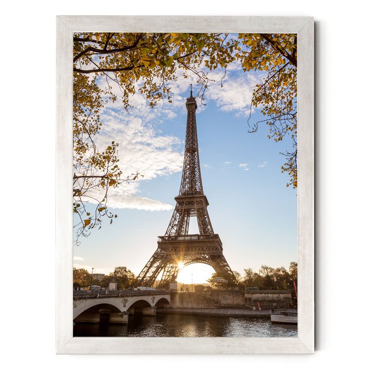 the Eiffel Tower in Paris, France, with a bridge and trees in the foreground, and the sun setting behind the tower.
