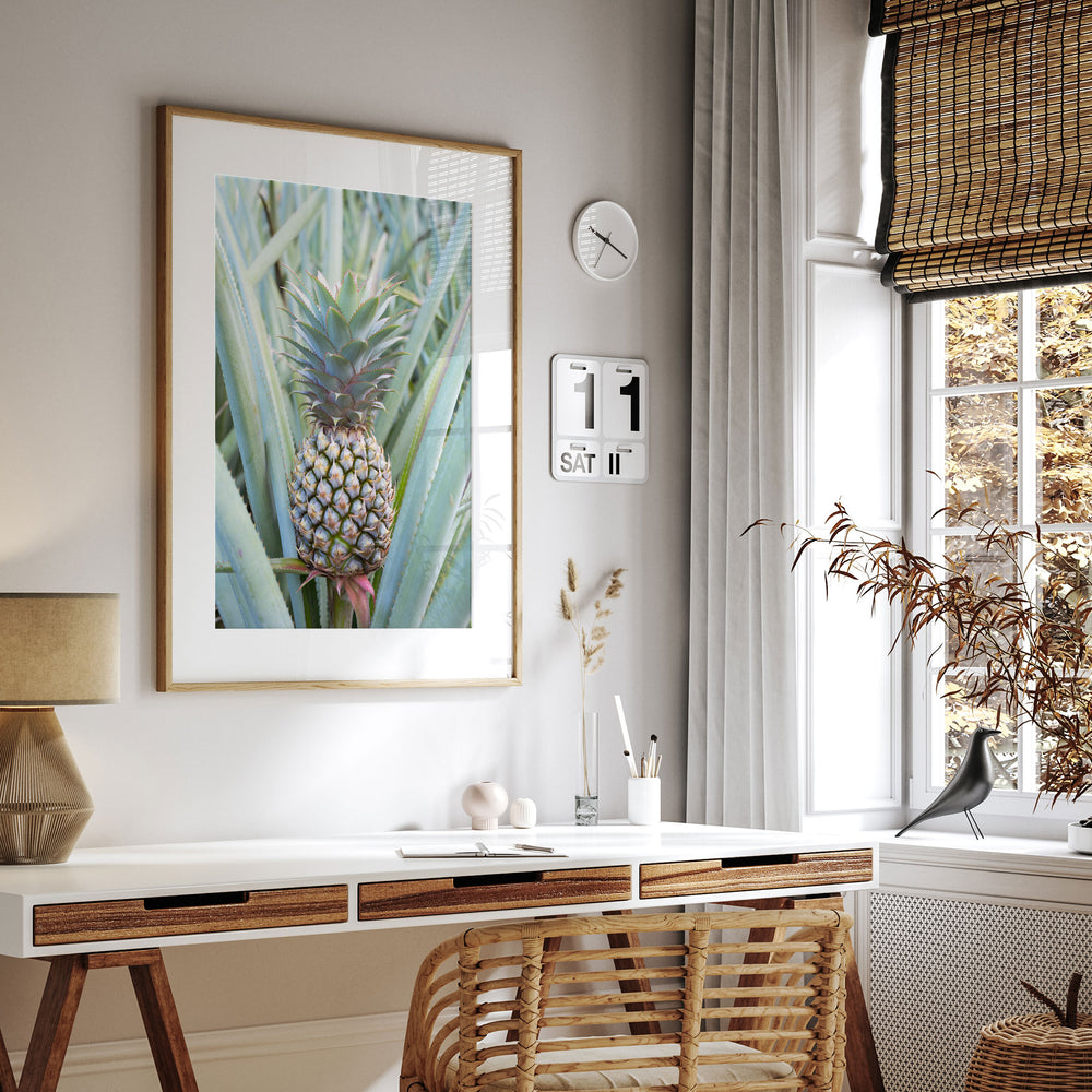 a modern and minimalist living space with a large framed print of a pineapple on the wall, a white desk with a lamp, a wicker chair, and a window with a bamboo shade.