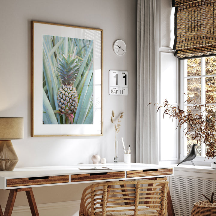 a modern and minimalist living space with a large framed print of a pineapple on the wall, a white desk with a lamp, a wicker chair, and a window with a bamboo shade.