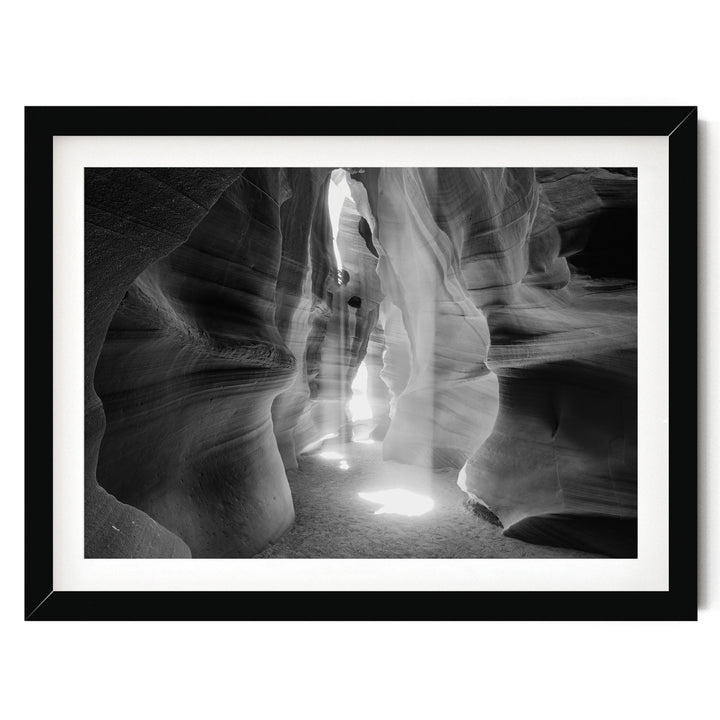 A black and white photograph of a narrow, winding path leading through a natural rock formation, with sunlight streaming through the opening and casting shadows on the walls.
