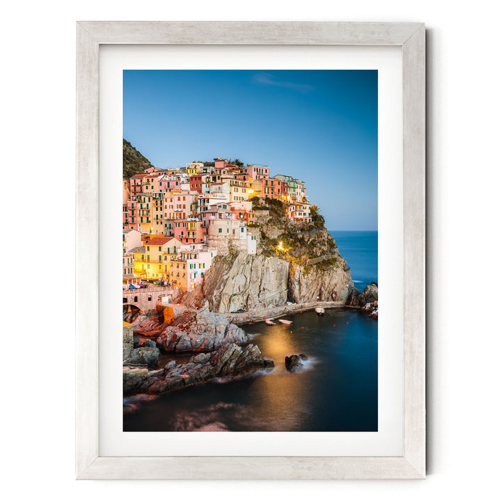 A picturesque coastal town nestled on a cliff, with its buildings illuminated by warm lighting against a backdrop of a clear blue sky and the ocean.
