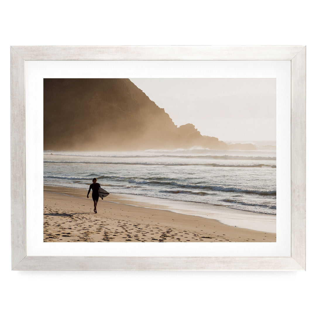 A person in a wetsuit walks along the beach, carrying a surfboard, with a rocky cliff and ocean in the background.