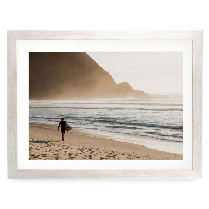 A person in a wetsuit walks along the beach, carrying a surfboard, with a rocky cliff and ocean in the background.