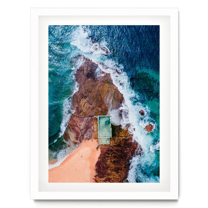 An aerial view of a beach with a swimming pool, surrounded by rocky cliffs and crashing waves.