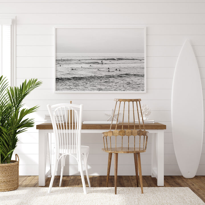 a serene and modern workspace with a wooden desk, two white chairs, a large framed picture of a beach scene, and a white surfboard leaning against the wall.