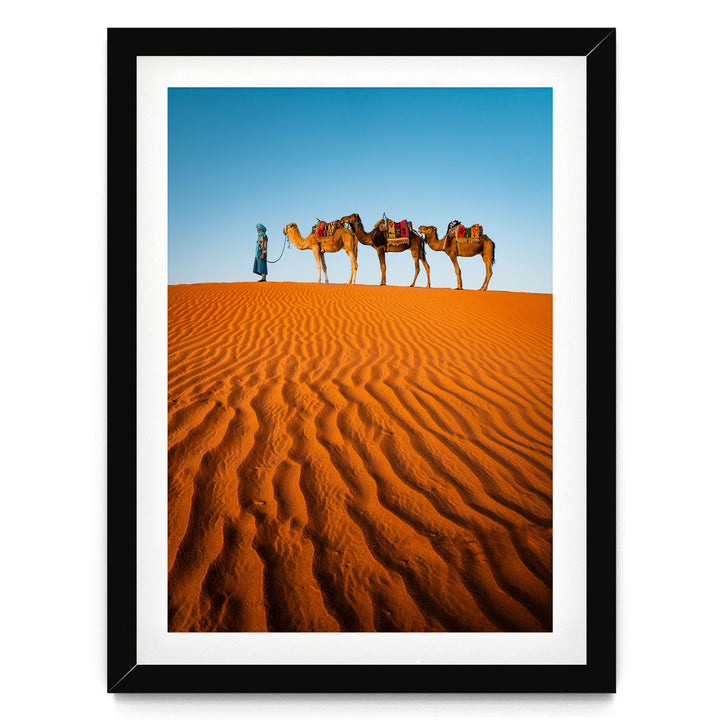 A person in blue attire leads three camels across a vast, undulating sand dune under a clear blue sky.