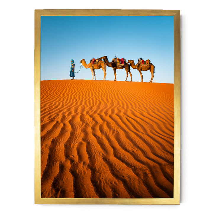A man in a blue robe leads three camels across a vast, undulating sand dune in a desert landscape.