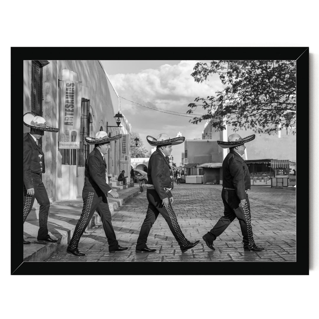 A group of men wearing sombreros and dark clothing are walking down a street, with a building and trees visible in the background.