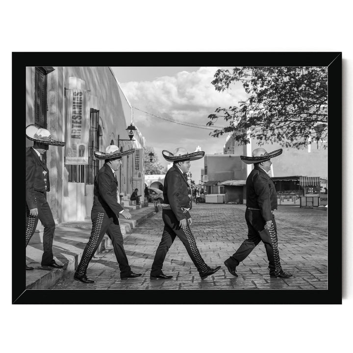 A group of men wearing sombreros and dark clothing are walking down a street, with a building and trees visible in the background.